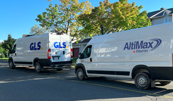 Une camionnette GLS suivie d'une camionnette AltiMax Courrier circulant dans une rue résidentielle.