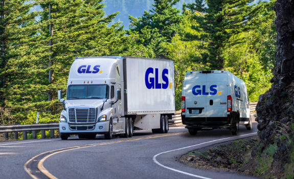 A GLS truck crosses a GLS parcel van on a road flanked by green trees under a clear sky..