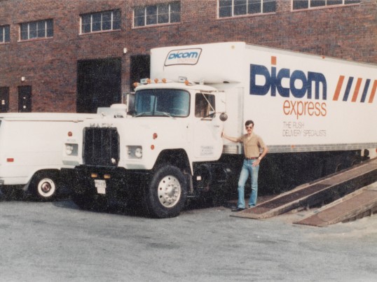 Man standing in front of a Dicom Express truck parked at a terminal in the 1960s.