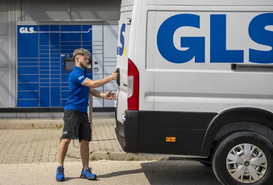 GLS courier in a blue polo shirt opening the rear door of a white GLS van, in front of GLS parcel lockers.