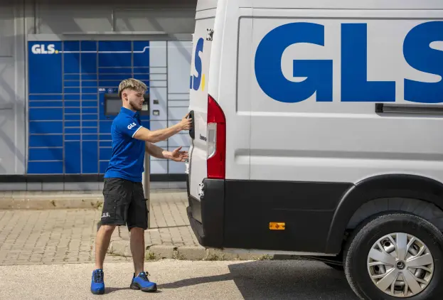GLS courier in a blue polo shirt opening the rear door of a white GLS van, in front of GLS parcel lockers.