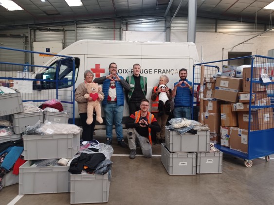 Volunteers pose in front of a French Red Cross van, surrounded by boxes of clothing, parcels, and stuffed animals collected for a solidarity action.