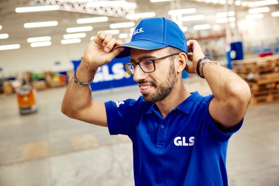 Smiling GLS employee in a blue uniform in a logistics warehouse, adjusting his cap.