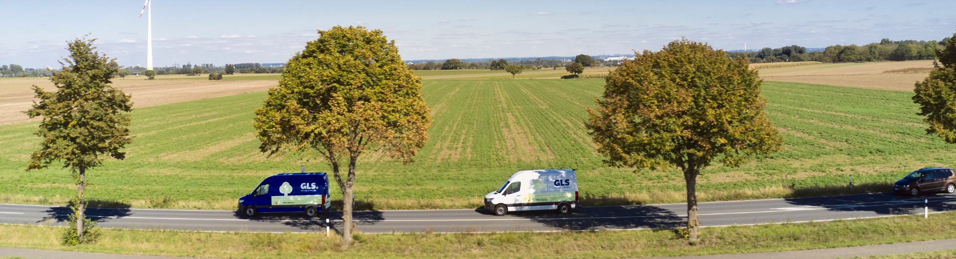 Two GLS delivery vans on a country road, with green fields and wind turbines on the horizon.