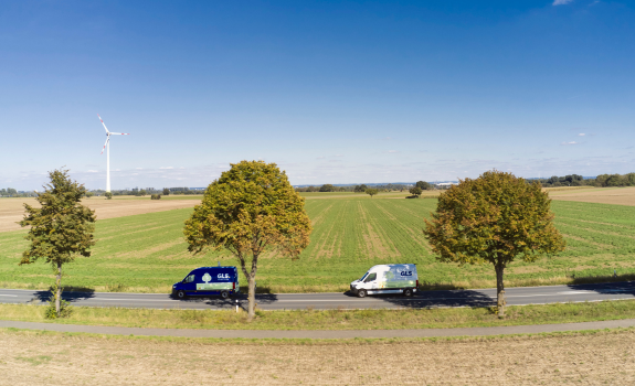 Two GLS delivery vans on a country road, with green fields and wind turbines on the horizon.