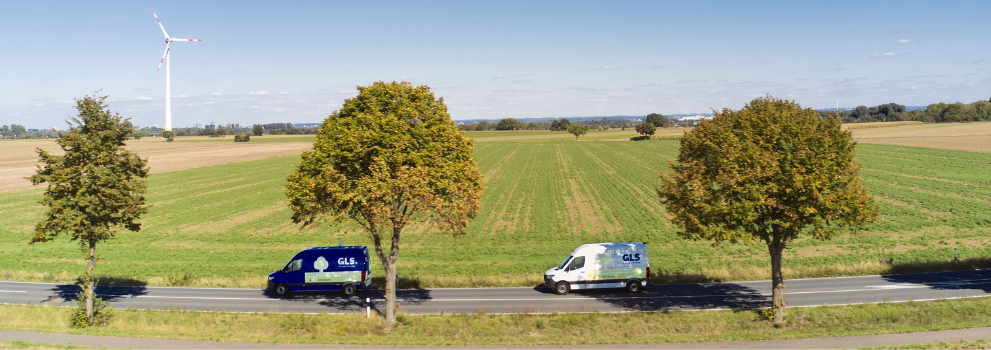 Two GLS delivery vans on a country road, with green fields and wind turbines on the horizon.