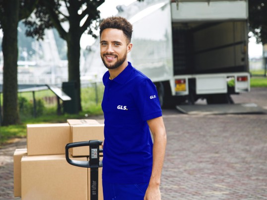 A GLS courier in a blue uniform stands beside stacked cardboard boxes and a hand truck in front of an open delivery truck.
