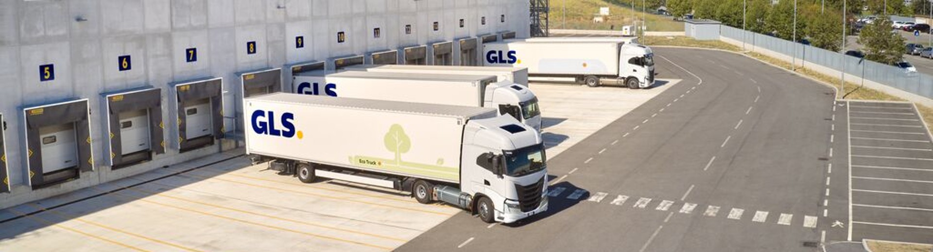 A large GLS logistics warehouse with loading dock bays and parked delivery trucks, seen from above beside paved roads and grassy areas.