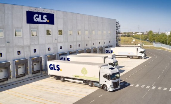 A large GLS logistics warehouse with loading dock bays and parked delivery trucks, seen from above beside paved roads and grassy areas.