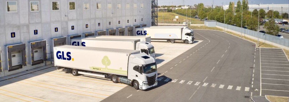 A large GLS logistics warehouse with loading dock bays and parked delivery trucks, seen from above beside paved roads and grassy areas.