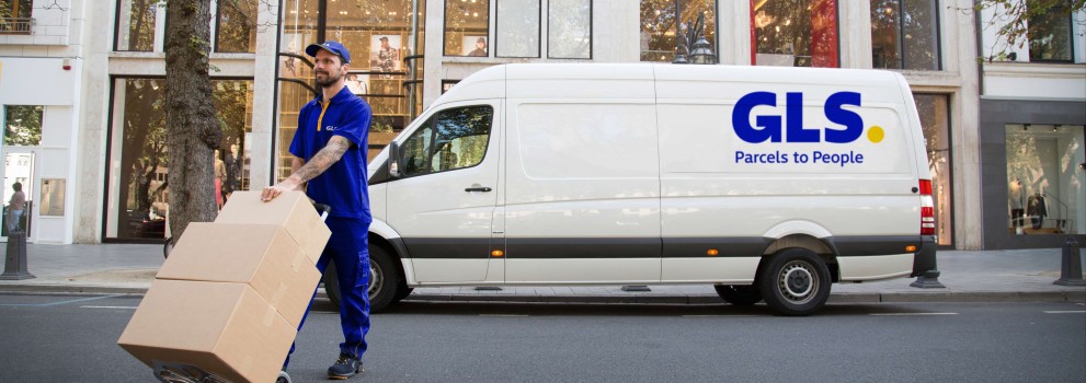 A GLS driver pushes packages on a dolly in front of a GLS van on a city street.